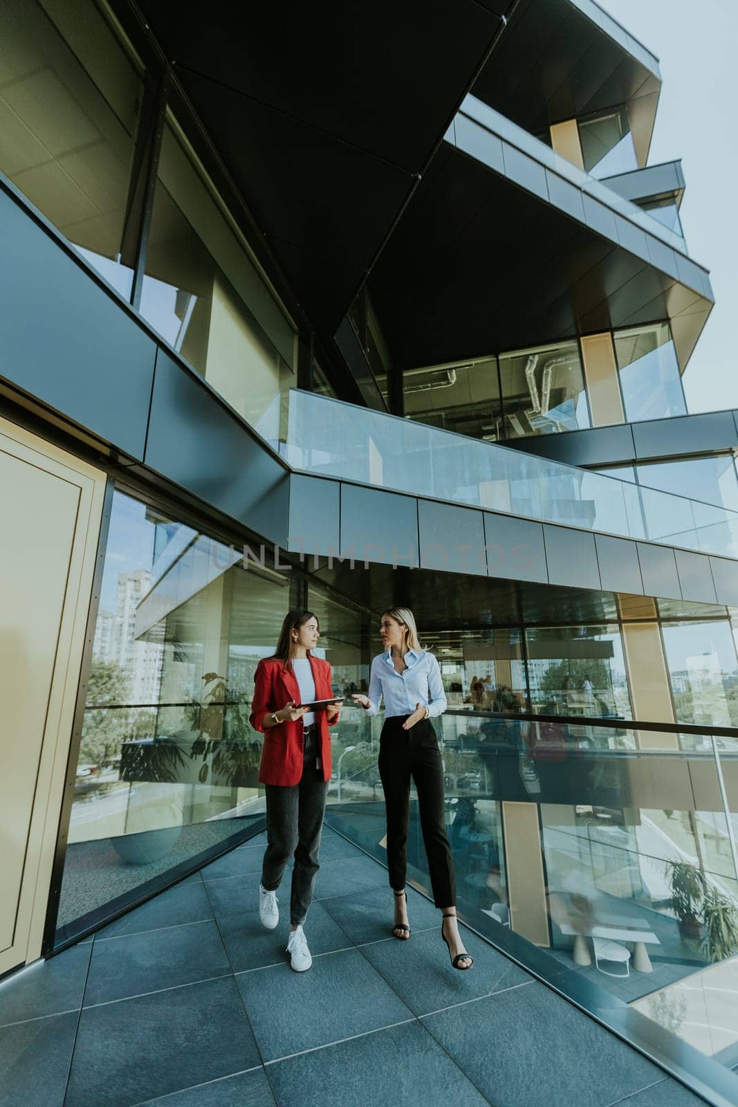 Two professional women engage in a thoughtful discussion while reviewing a tablet outside a sleek, contemporary office building during the daytime.