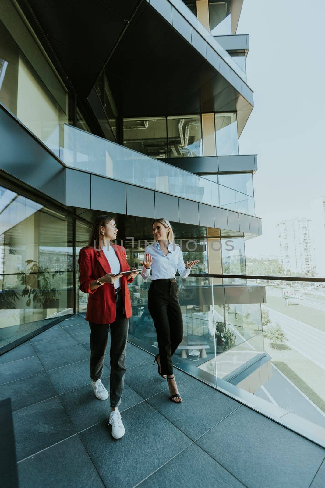 Two professional women engage in a thoughtful discussion while reviewing a tablet outside a sleek, contemporary office building during the daytime.