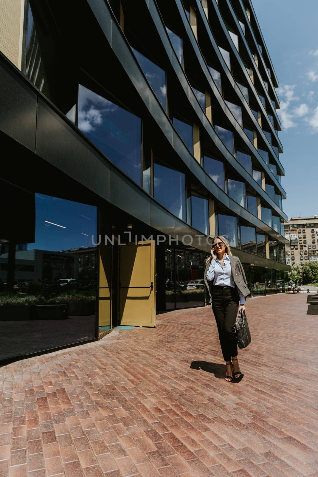 A woman in business attire walks along a brick path outside a sleek, architectural office building under a clear blue sky.