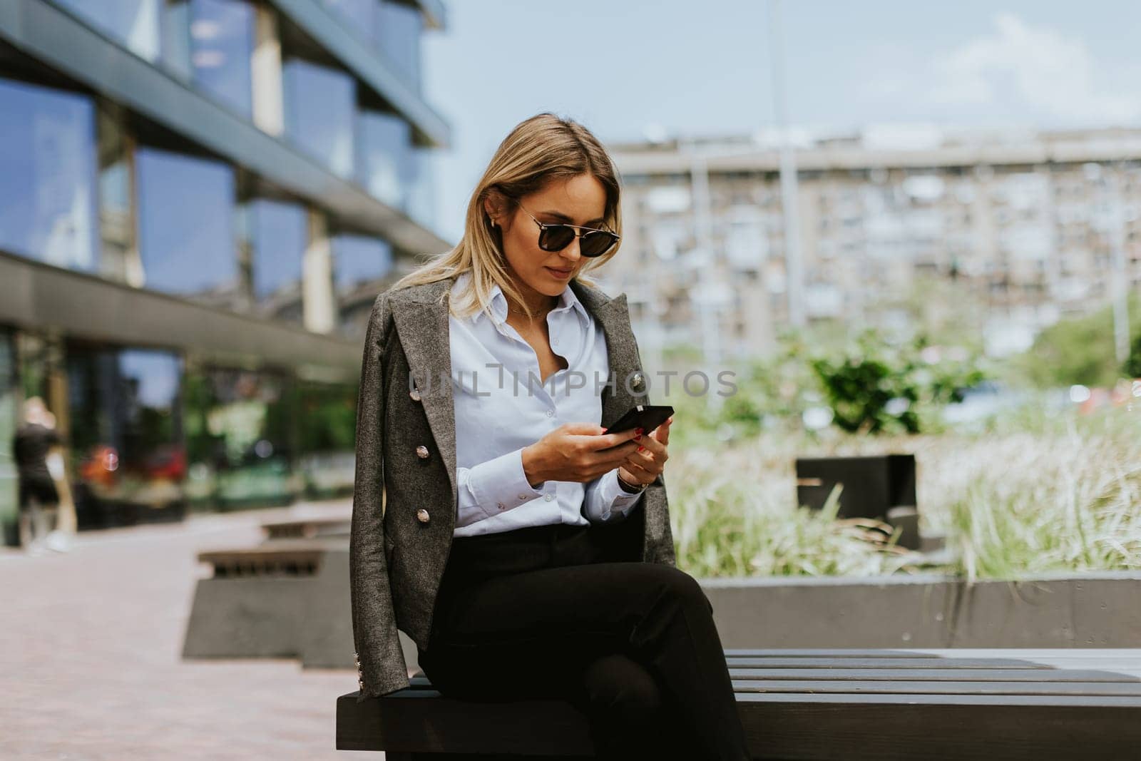 A woman with long dark hair sits on a bench, engrossed in her phone screen, her expression thoughtful and absorbed.