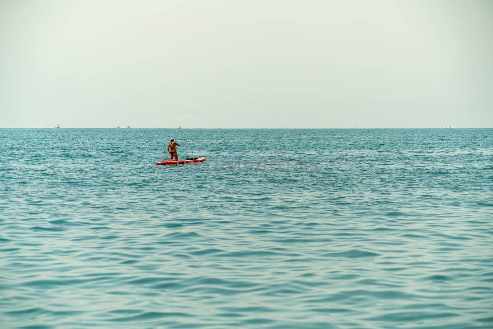 A person paddles on a red board across tranquil blue waters under a hazy sky. Nearby, small boats are visible in the distance, creating a serene atmosphere of relaxation.