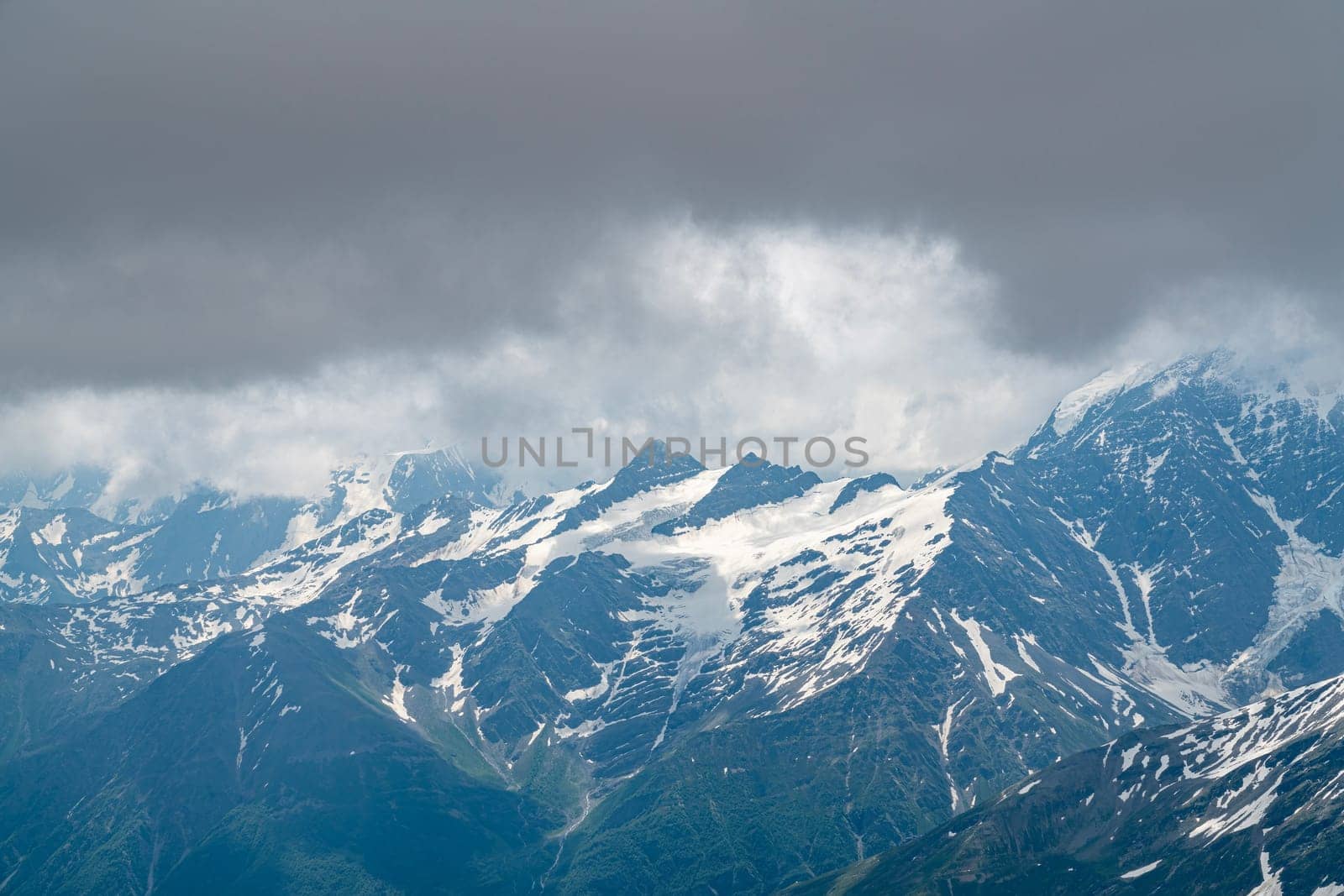 Snow-covered peaks rise dramatically against a cloudy sky. The landscape showcases rugged beauty, with patches of green highlighting the mountain slopes in midday light.