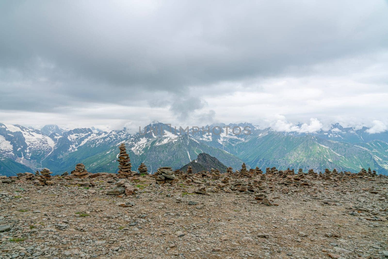 A series of stone cairns are arranged on rocky terrain, with towering snow-capped mountains and dramatic clouds in the distance. The landscape showcases the beauty of nature's formations.