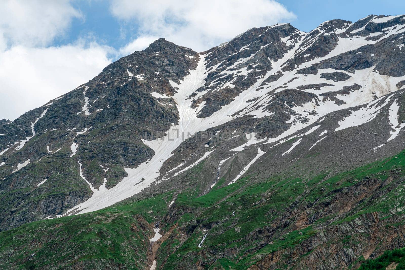 A majestic mountain rises with patches of snow and vibrant green slopes under a clear blue sky. The beauty of nature is highlighted in this serene alpine setting.