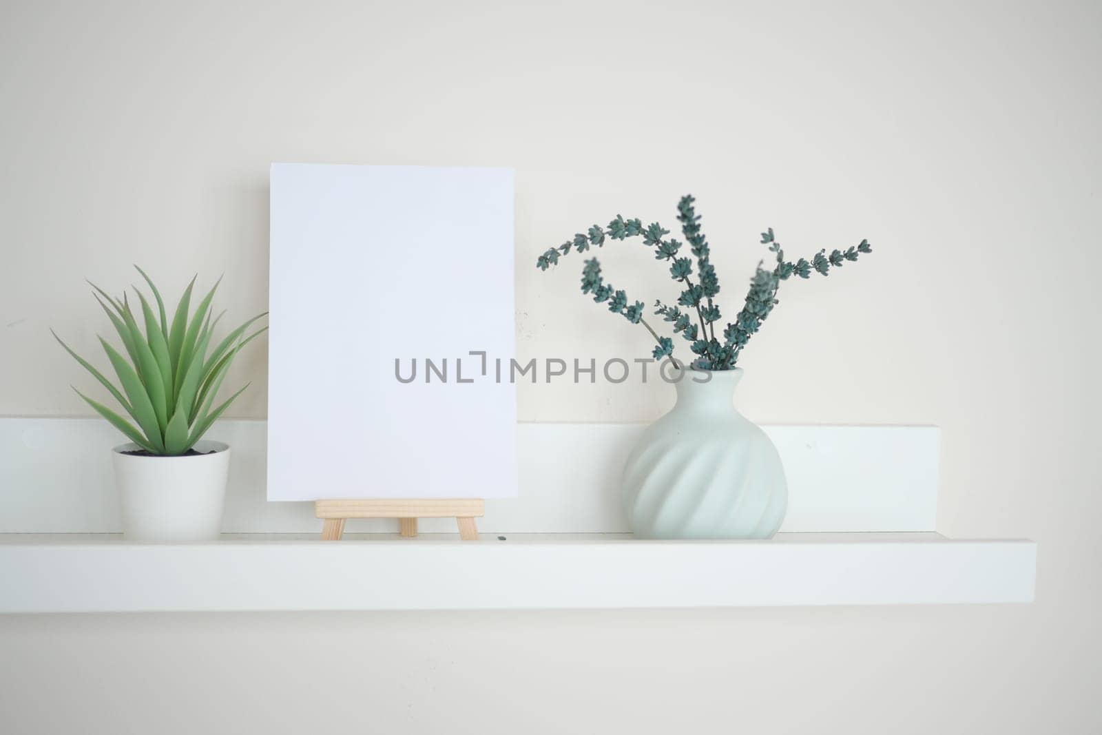 A simple shelf features a blank canvas, a plant, and a vase of greenery.
