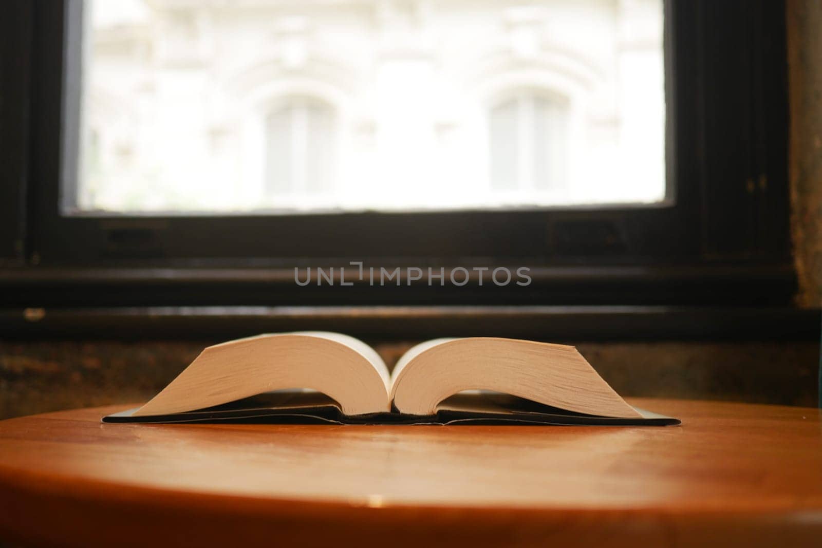 An open book lays on a round wooden table beside a window, inviting readers.