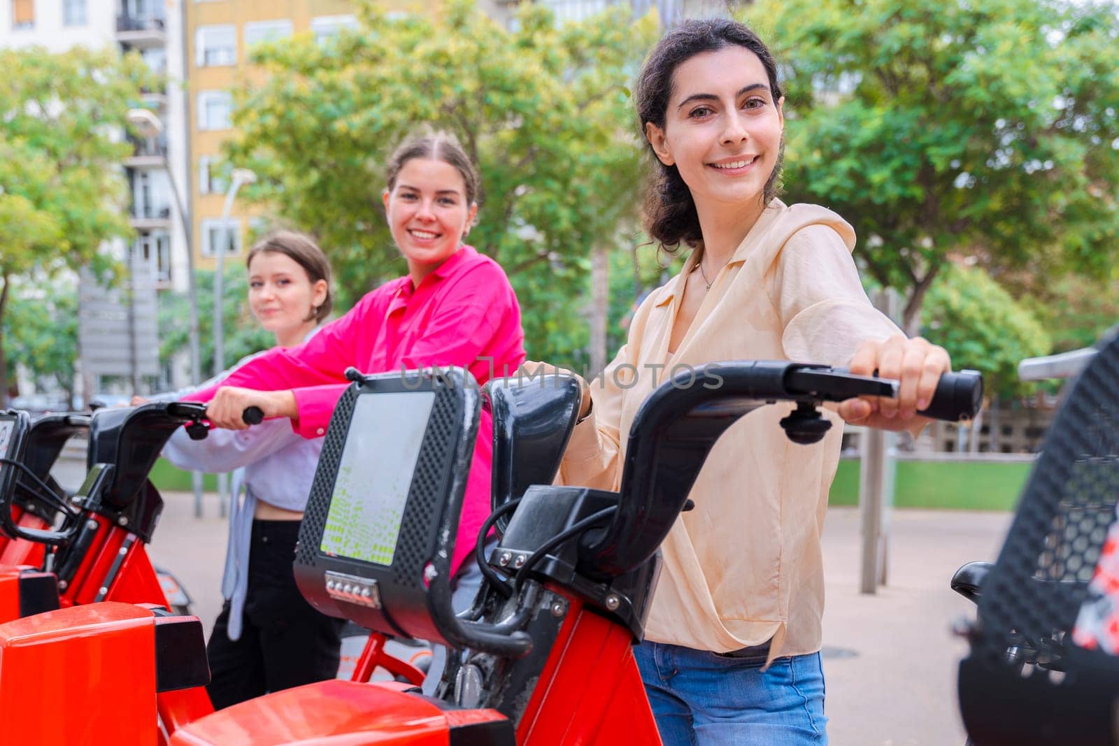 Happy young women renting bicycles in an urban park, smiling and preparing for a fun ride. Dressed casually, they enjoy a lively outdoor setting surrounded by trees and modern city buildings.