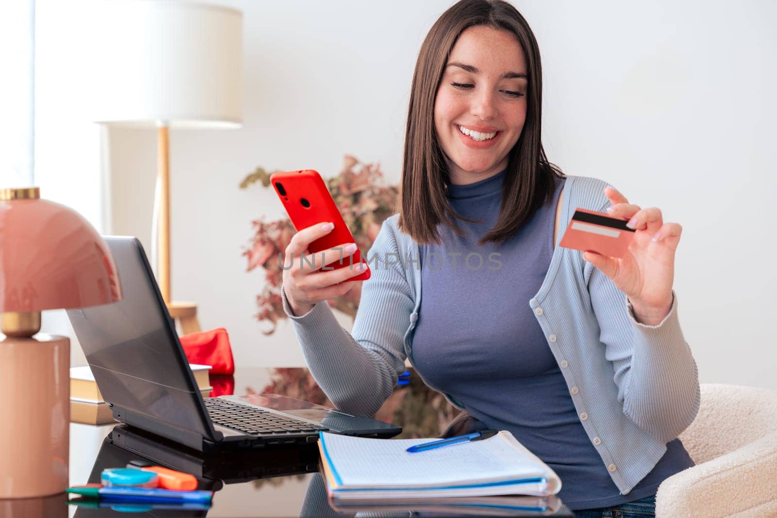 Caucasian student making an online payment with a credit card while using a phone at home. Modern workspace with books, stationery, and stylish decor. E-commerce, digital transaction, and finance.