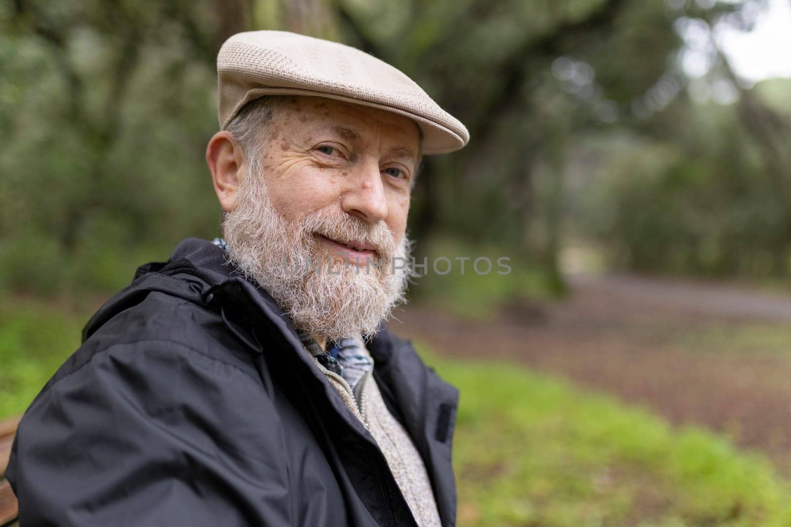 Portrait of a serene senior man with a white beard wearing a flat cap, sitting in a park and enjoying the peaceful atmosphere