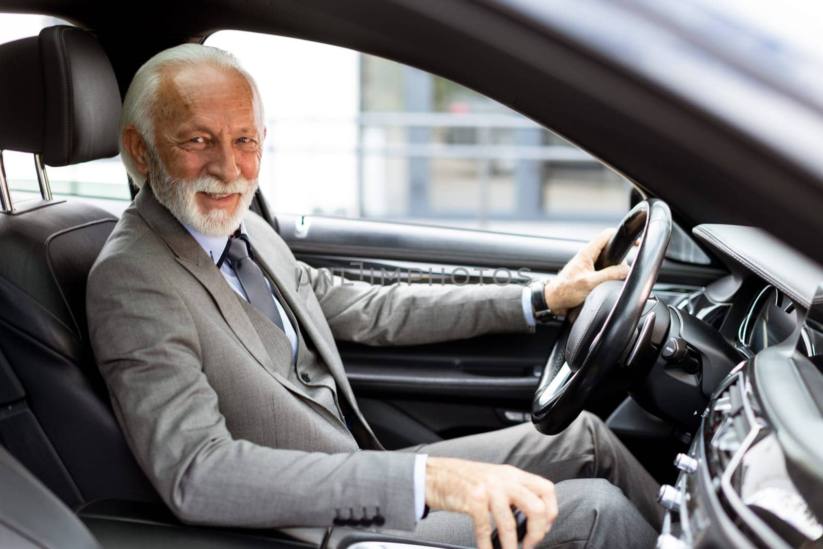 An elderly gentleman, dressed in a suit, confidently drives a modern car through a bustling city street on a bright, sunny day