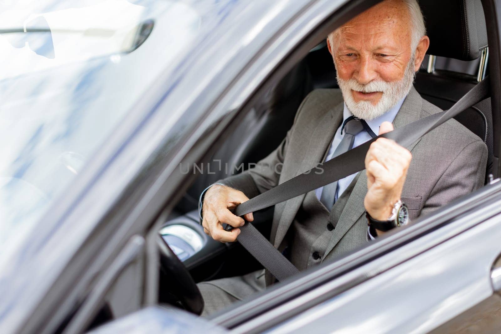 Elderly man with white hair and a beard is sitting in a modern car, fastening his seatbelt and smiling on a sunny afternoon drive