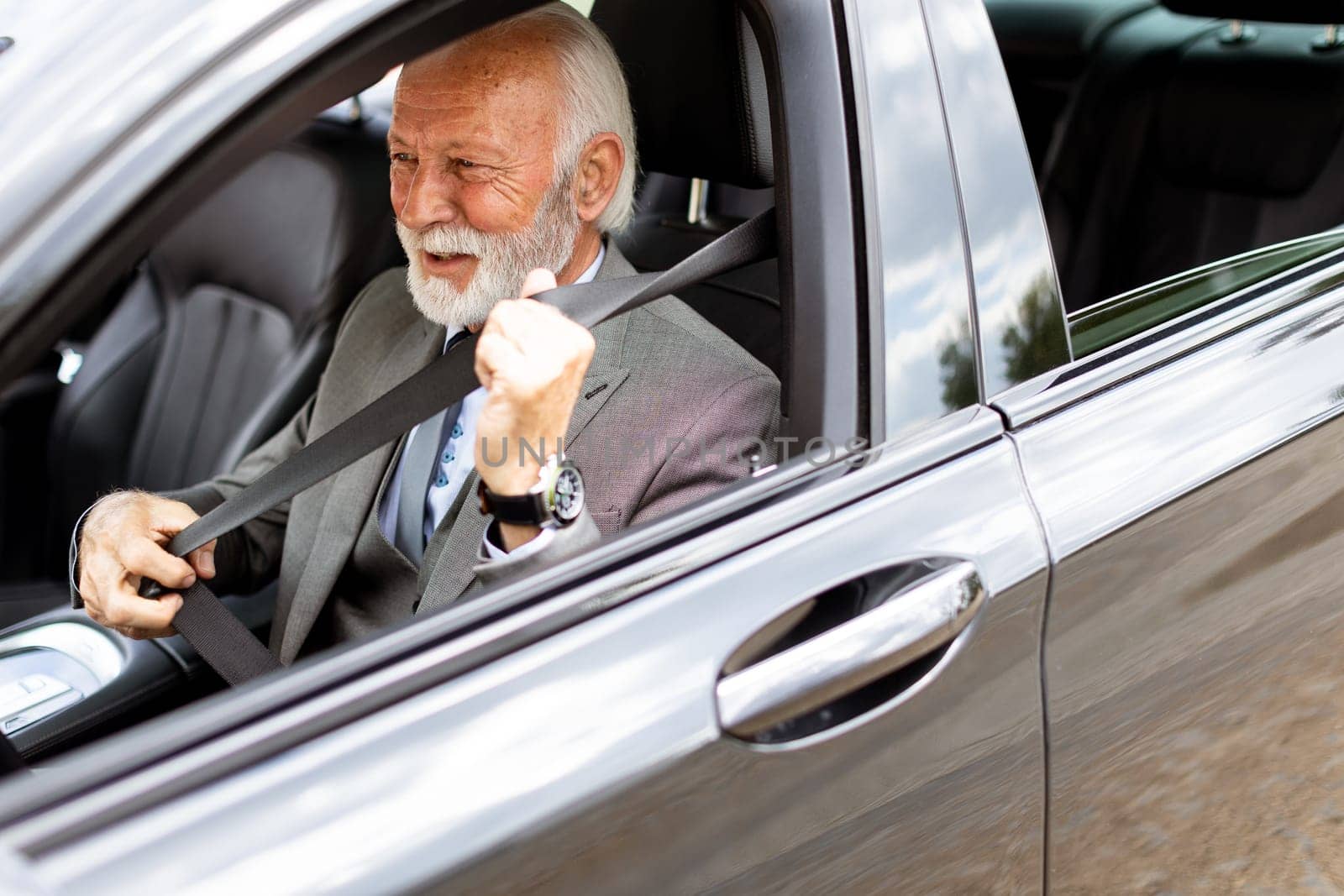 Elderly man with white hair and a beard is sitting in a modern car, fastening his seatbelt and smiling on a sunny afternoon drive