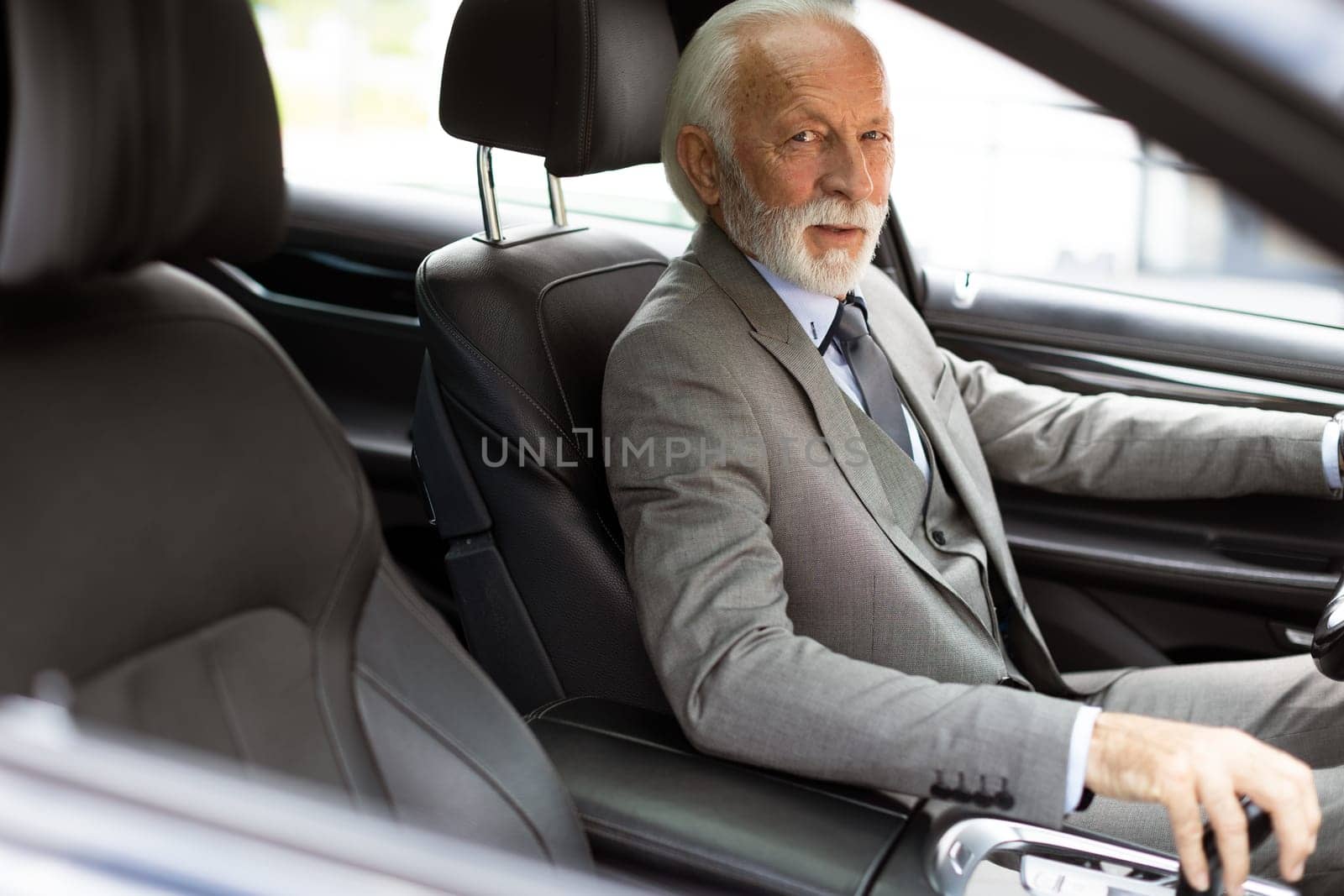 An elderly gentleman, dressed in a suit, confidently drives a modern car through a bustling city street on a bright, sunny day