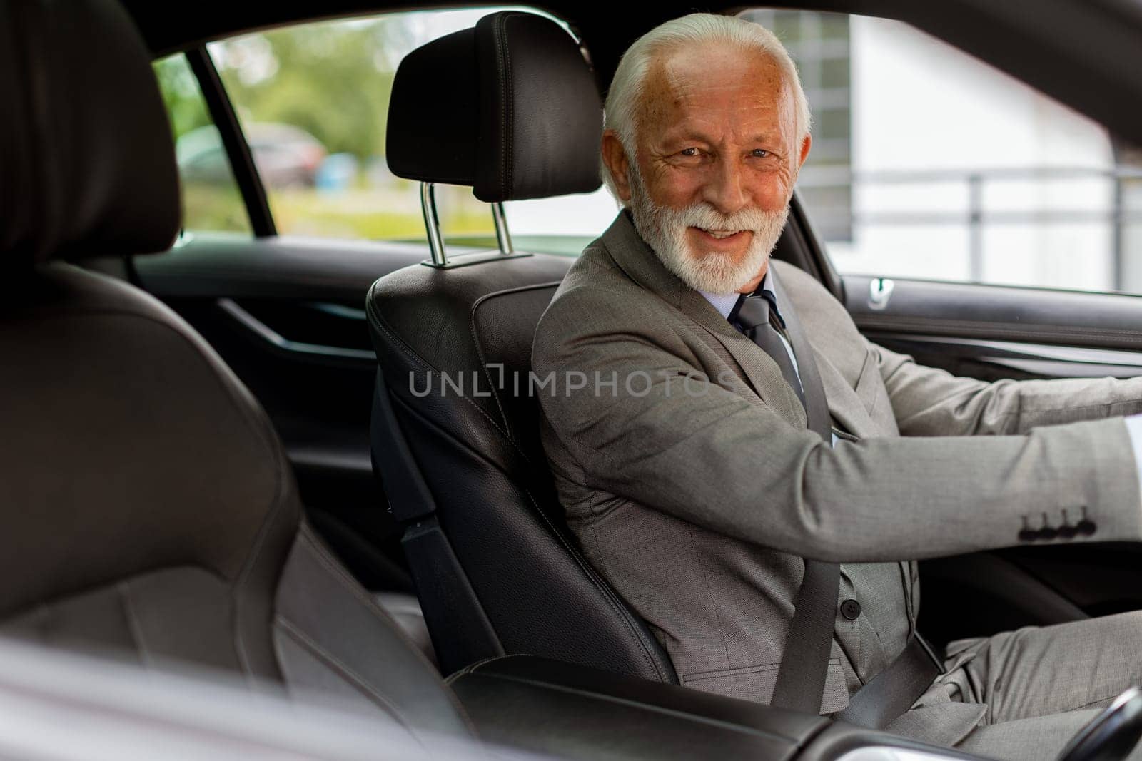 An elderly gentleman, dressed in a suit, confidently drives a modern car through a bustling city street on a bright, sunny day