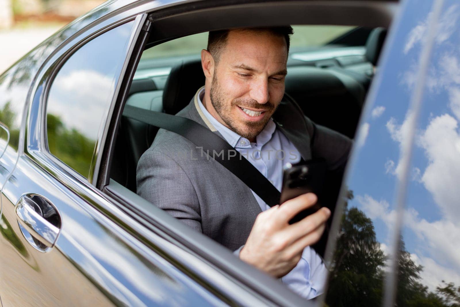 Smiling man in business attire checks his phone while seated in the backseat of a moving car