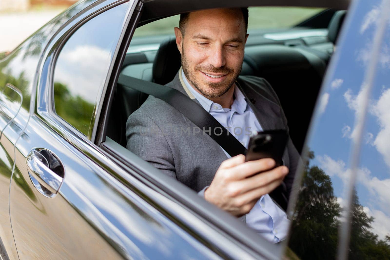 Smiling man in business attire checks his phone while seated in the backseat of a moving car