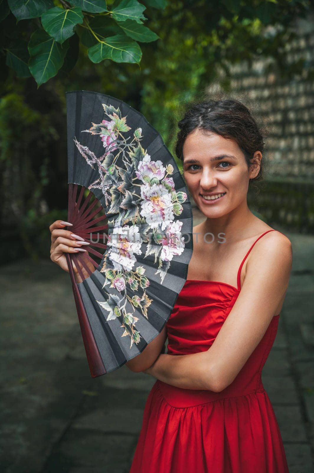 beautiful happy young woman in red dress with a fan in the garden. Tourism and travel in Asia