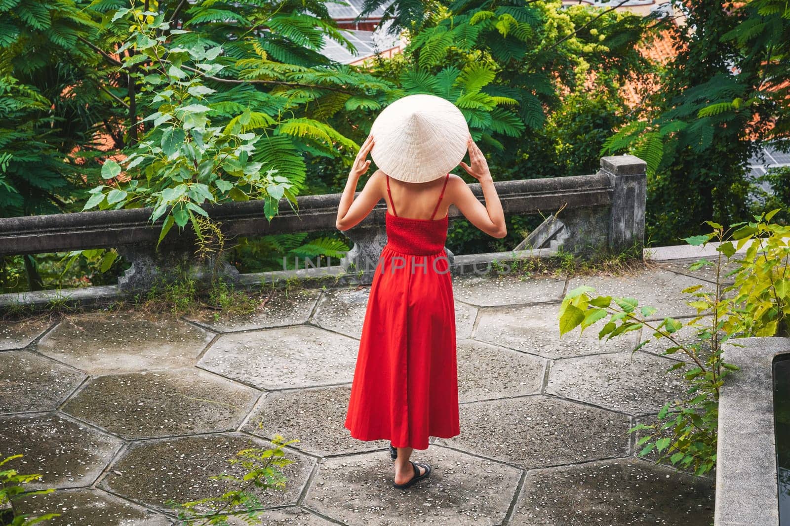 back of beautiful girl tourist traveler in a Vietnamese non la hat in red dress in a garden in Vietnam. Tourism and travel in Asia