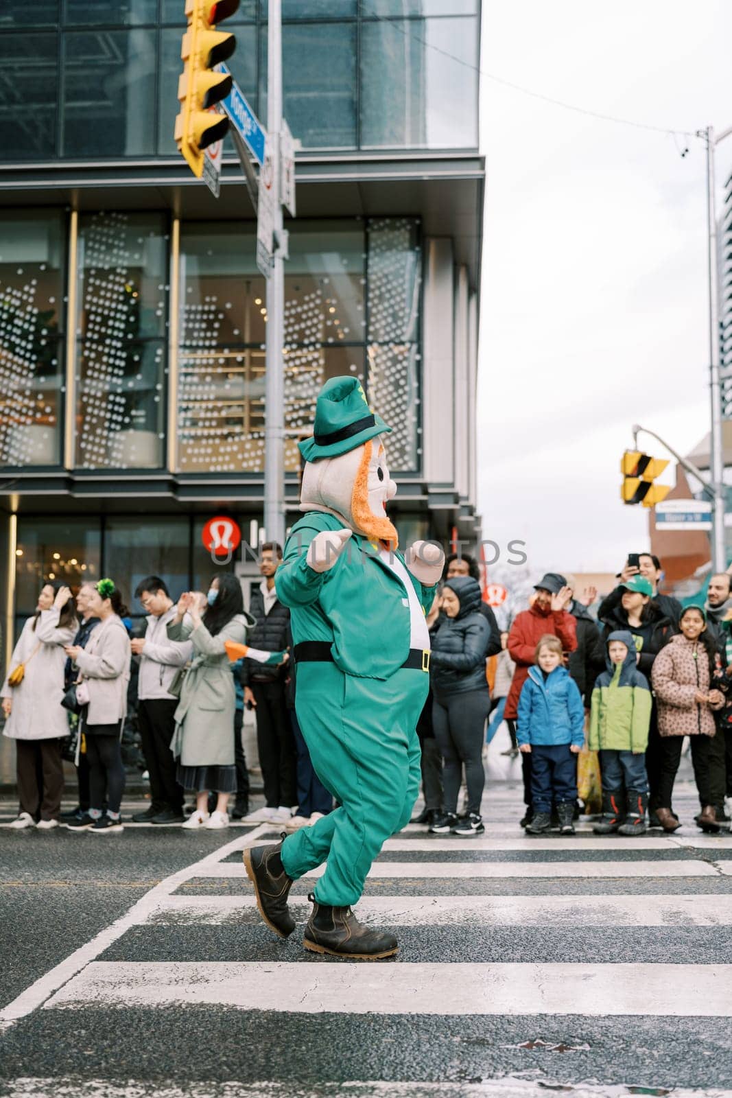 A lively leprechaun walks through a festive parade crowd on the street.