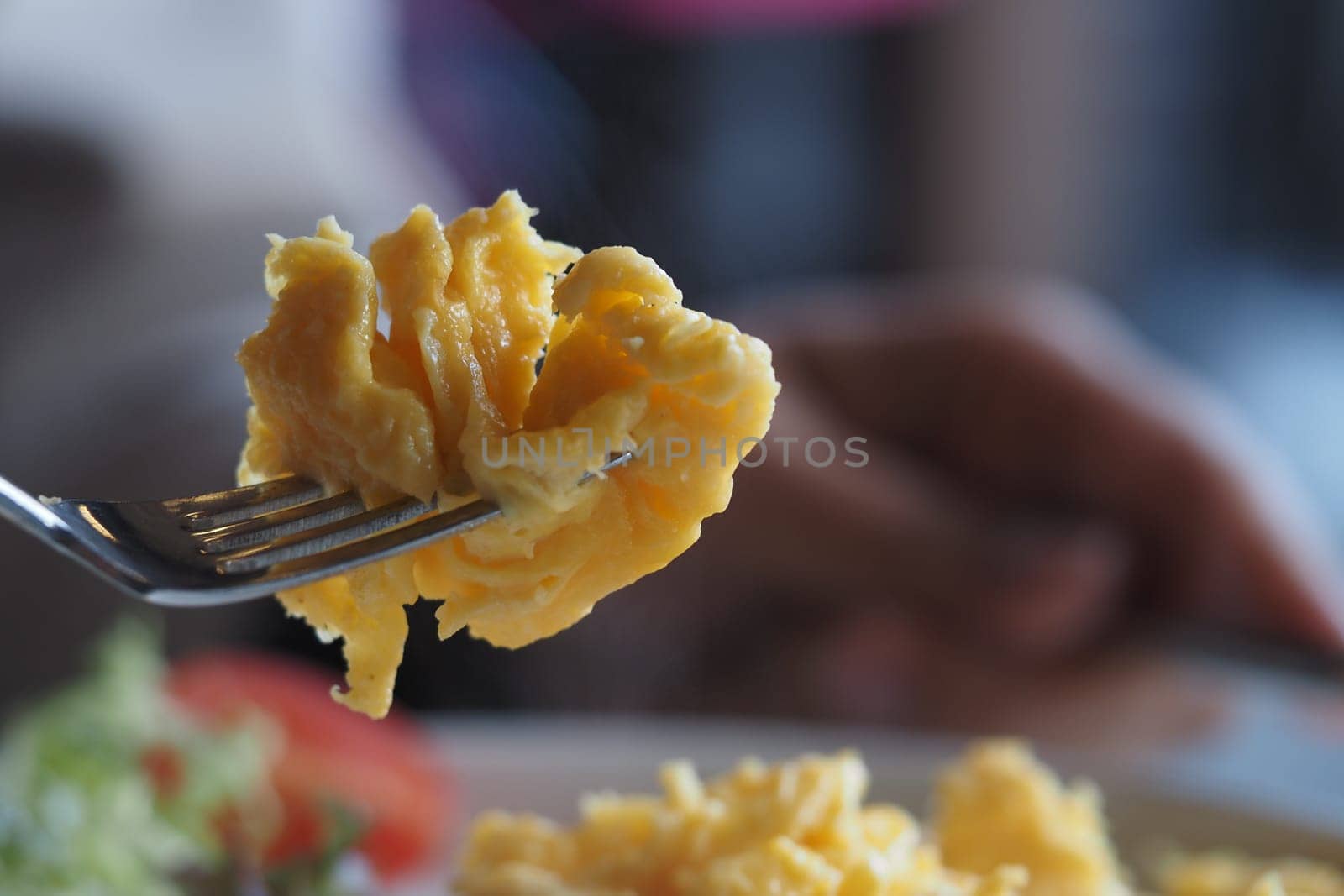 Fork holding fluffy scrambled eggs near a breakfast dish with vegetables.
