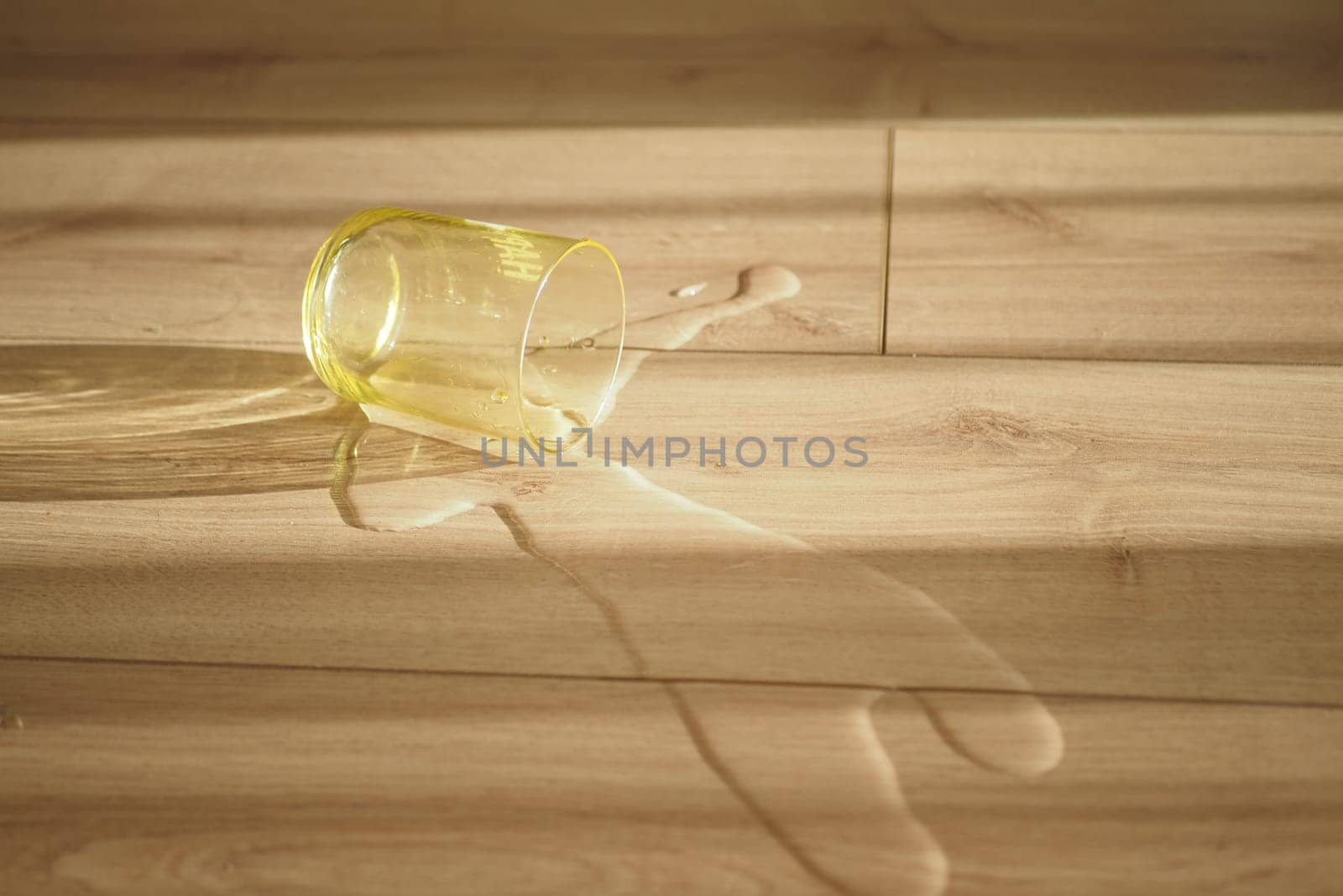 A yellow glass lies on a wooden floor with water spilling out, cast in sunlight.