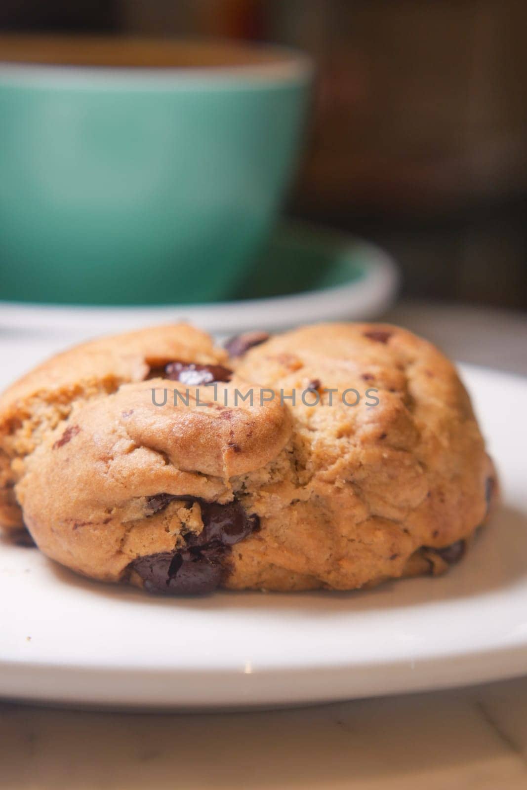 A chocolate chip cookie rests on a white plate beside a coffee cup.