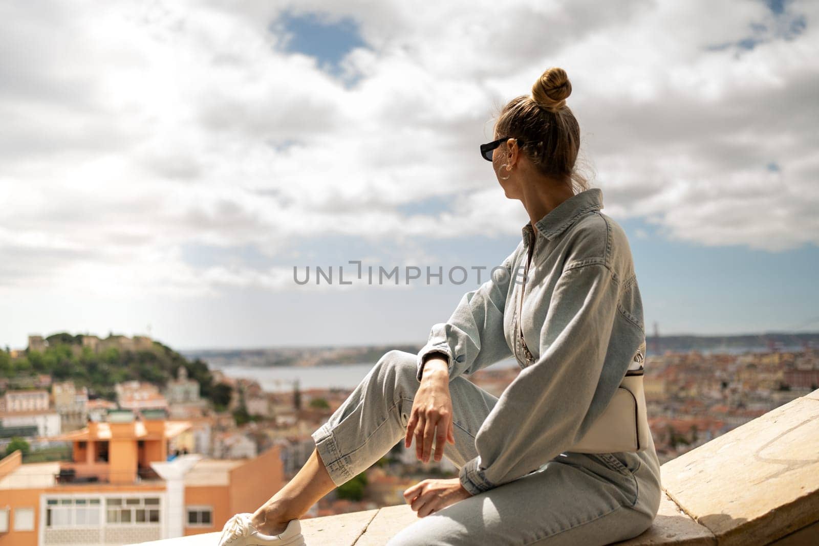 Young woman sitting on wall, enjoying the view of old town Lisbon. Tourist at Miradouro da Graca viewpoint, admiring scenic cityscape and architecture