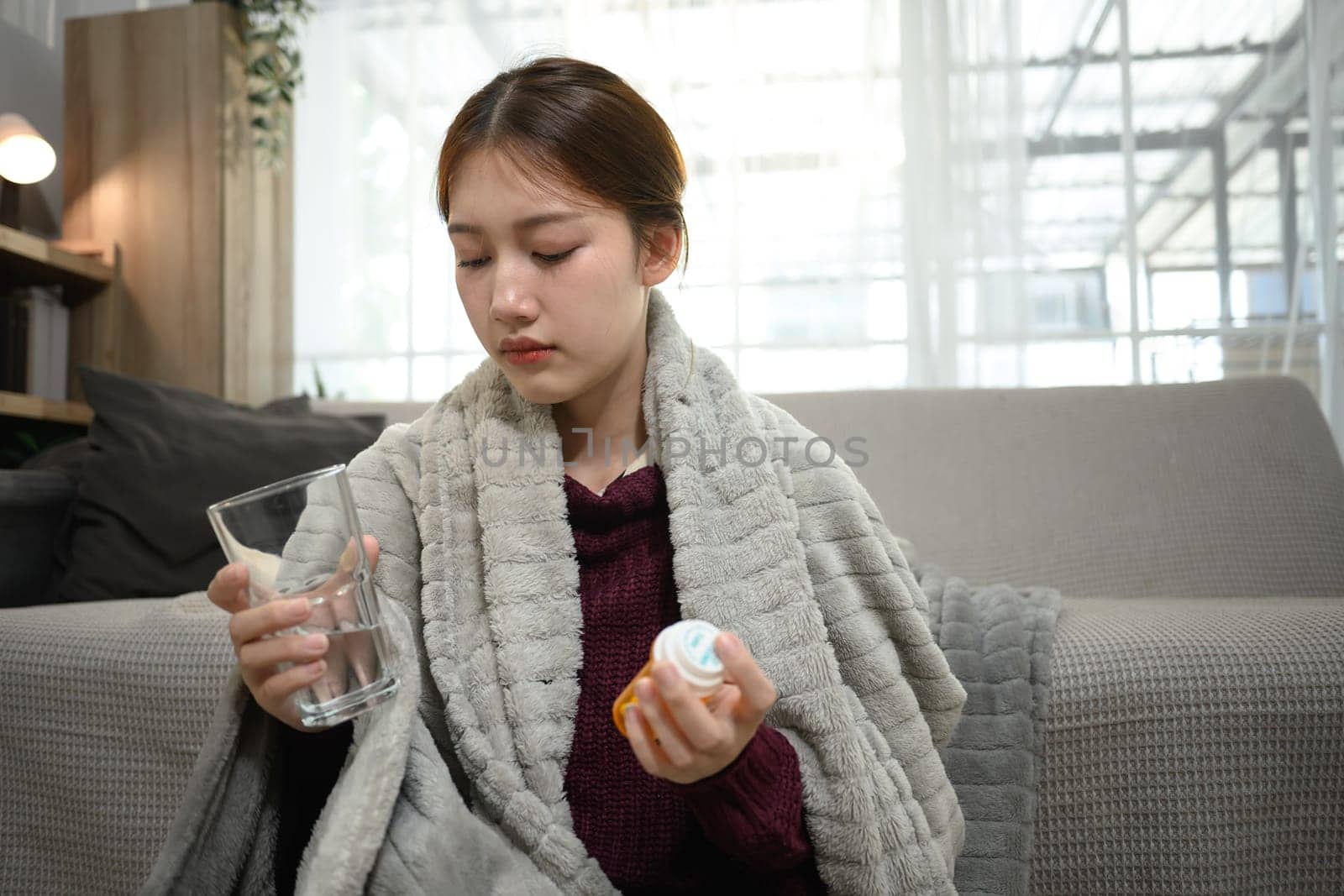Sick young woman wrapped in warm blanket, holding a glass of water and a pill bottle.