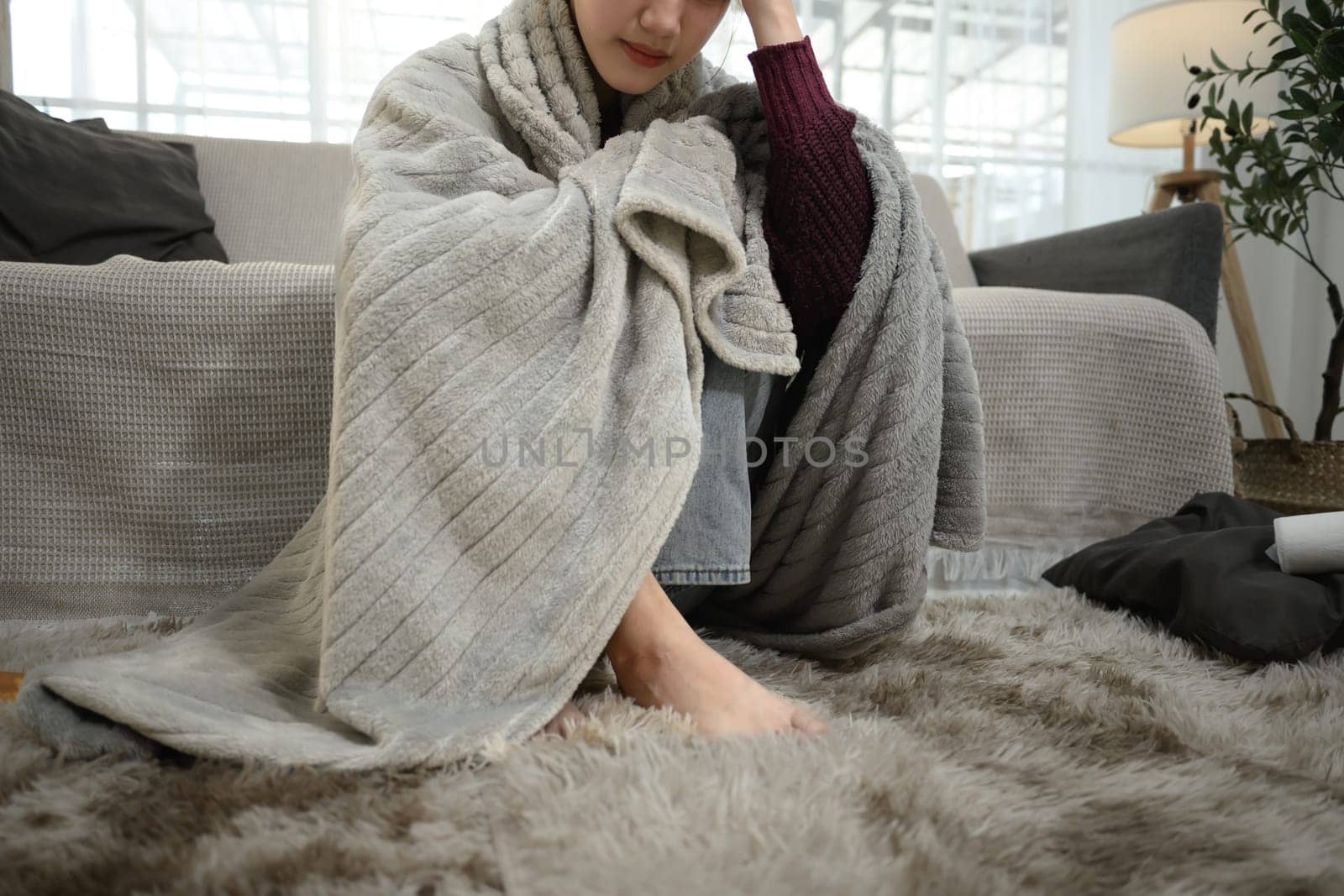 Unwell woman wrapped in warm blanket, holding her head in discomfort. Illness and home recovery concept.