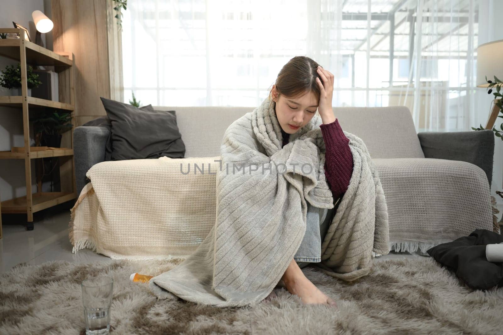 Unwell woman wrapped in warm blanket, holding her head in discomfort. Illness and home recovery concept.