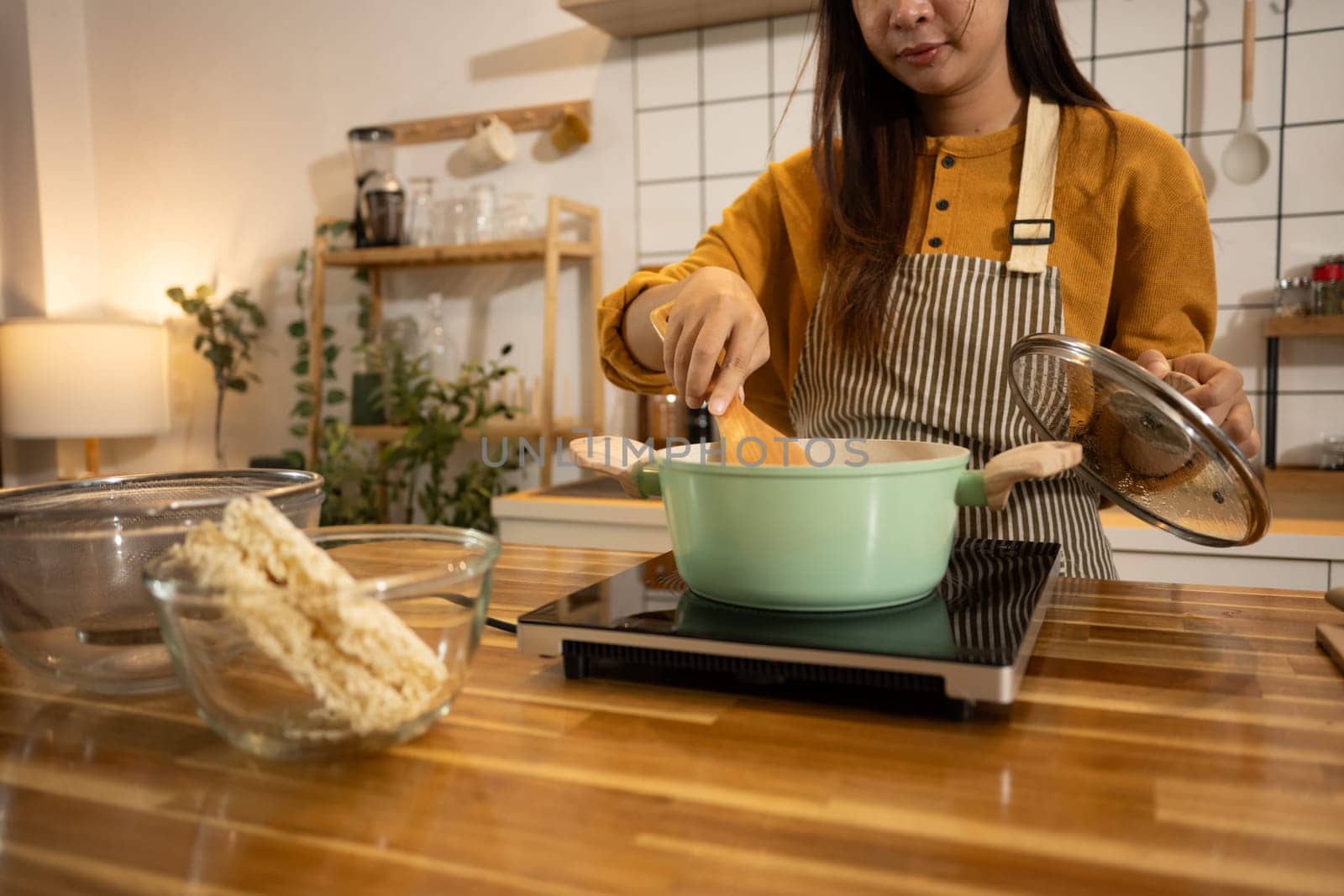 Young asian woman stirs pot on stove, preparing a homemade meal in a kitchen.