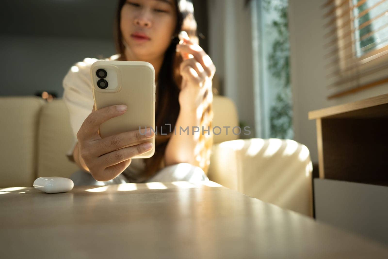 Young woman wearing a wireless earbud and using a smartphone in bright living room.