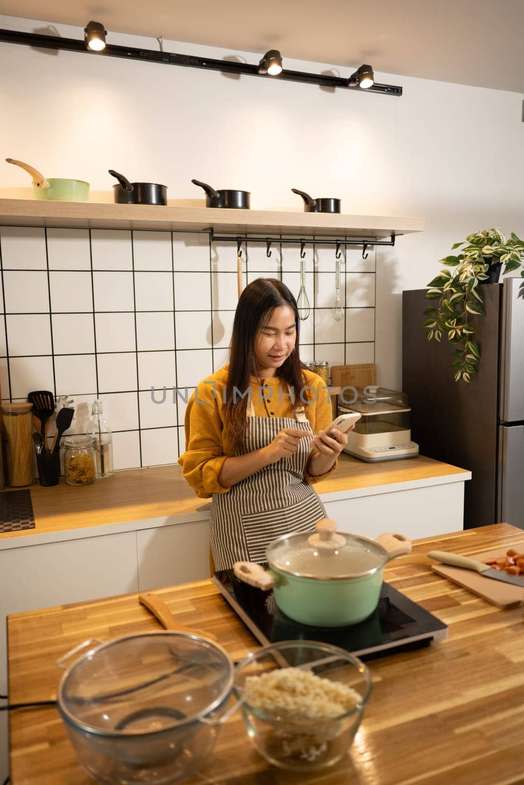 Smiling asian woman wearing an apron using smartphone while preparing meal in a kitchen.