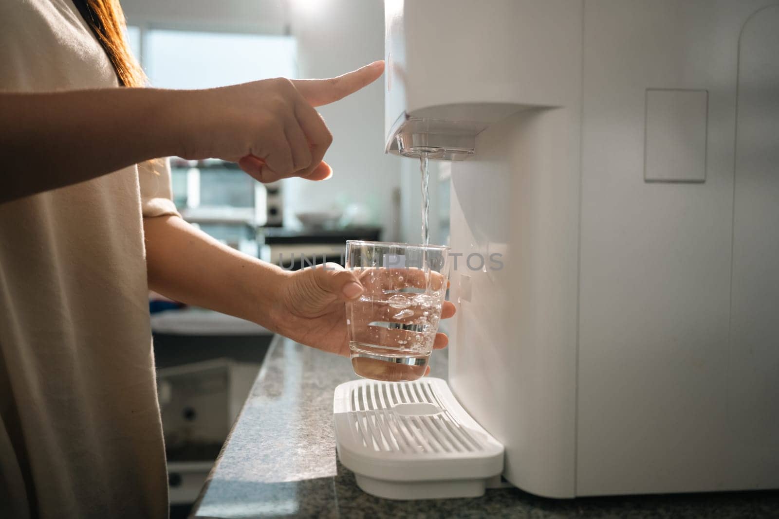 Woman using a modern water purifier in kitchen, filling a glass with fresh, purified water.