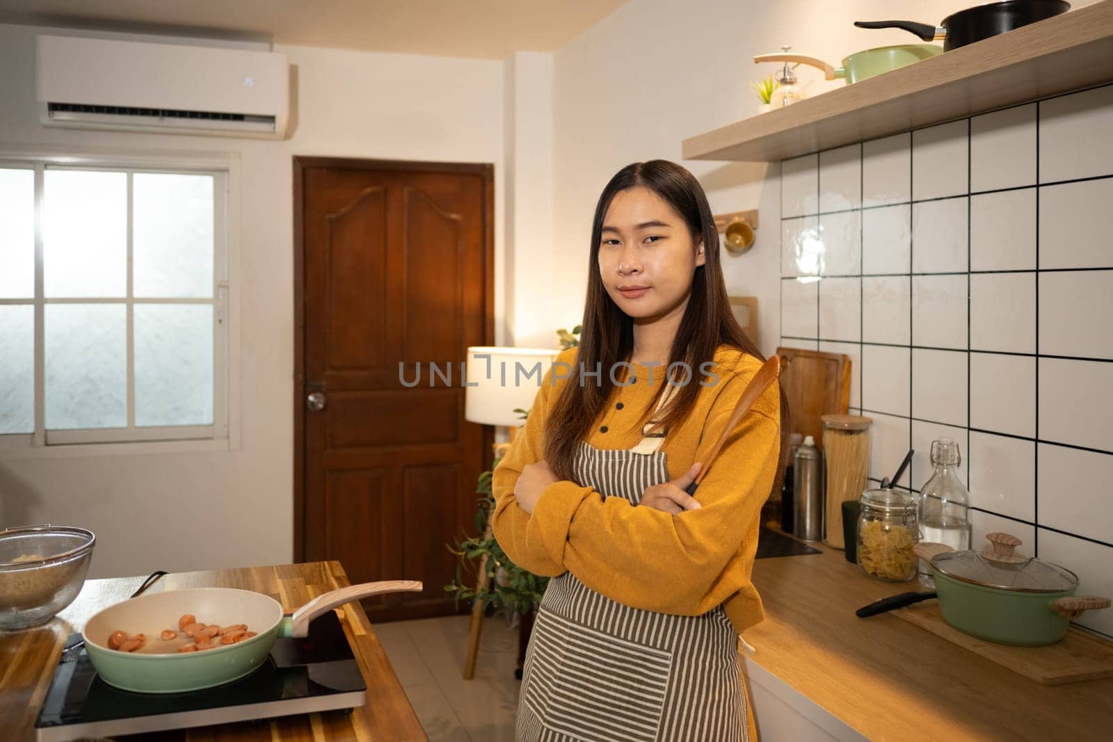 Proud young woman standing with arms crossed, holding a wooden spatula in a kitchen.