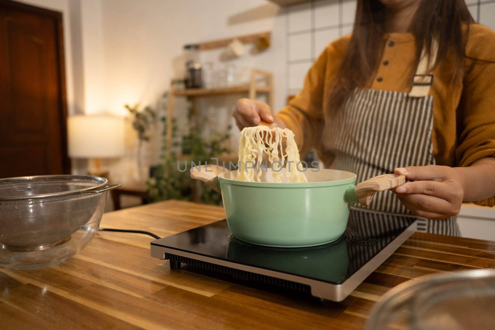 Close up woman lifts cooked noodles from a green pot on an induction stove.