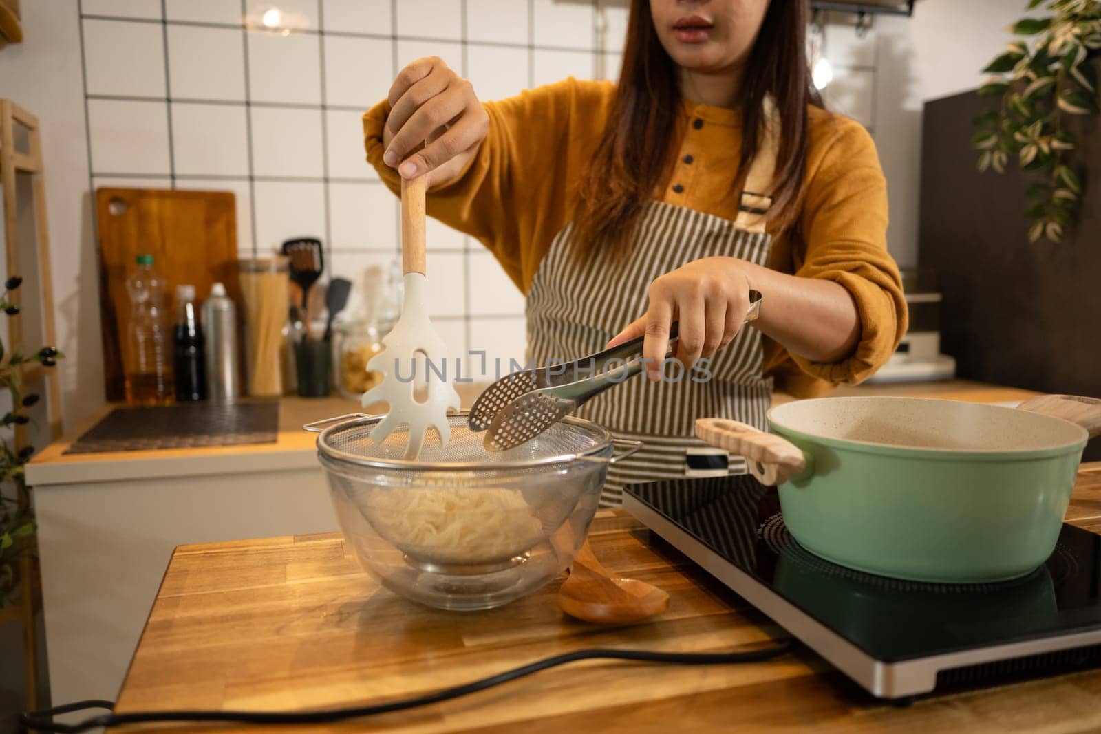 Woman wearing apron using kitchen utensils to transfer cooked noodles into a strainer.