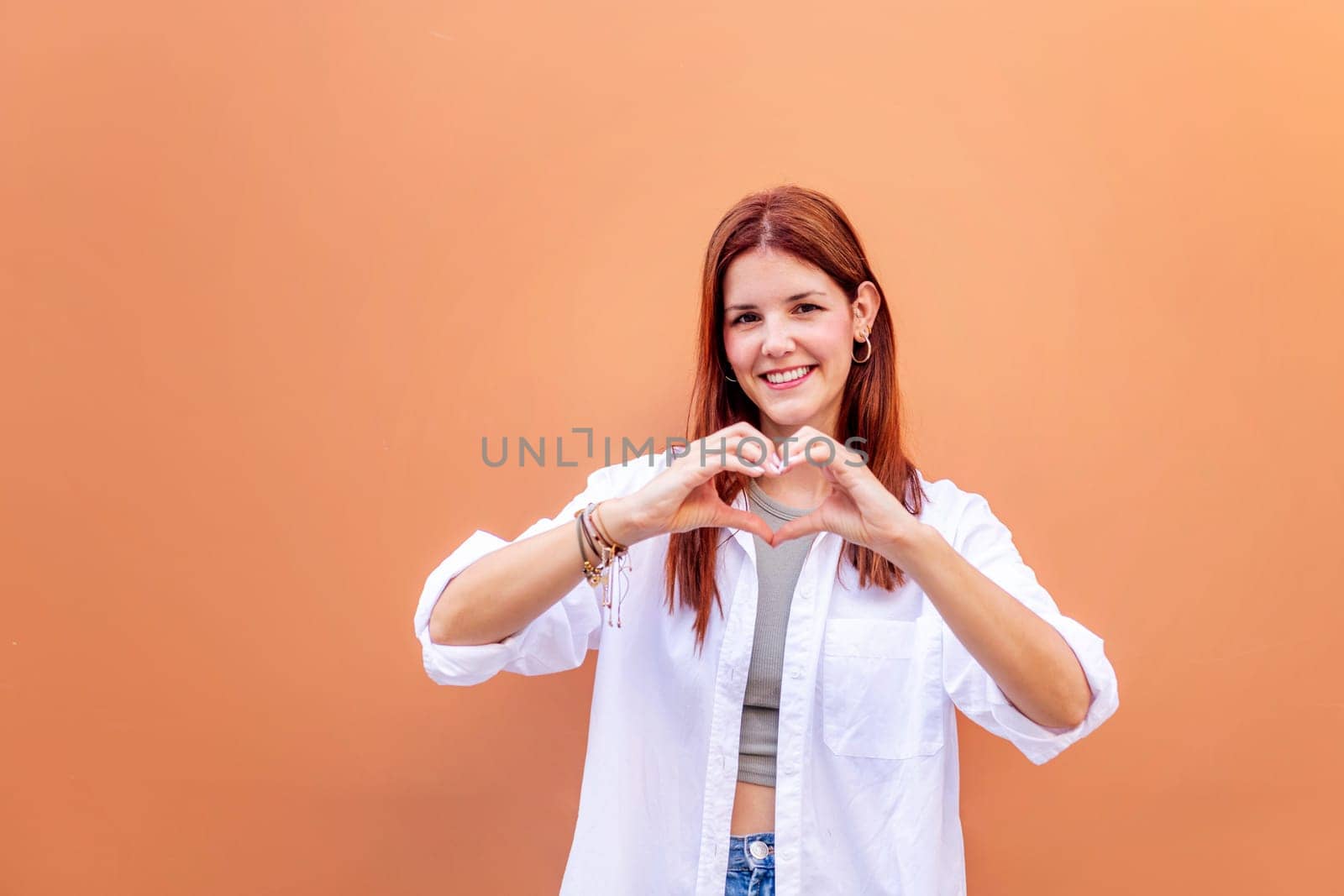 young woman standing in front of orange wall doing a heart shape with hands while looking at camera, she is wearing a white shirt, concept of love, copyspace for text