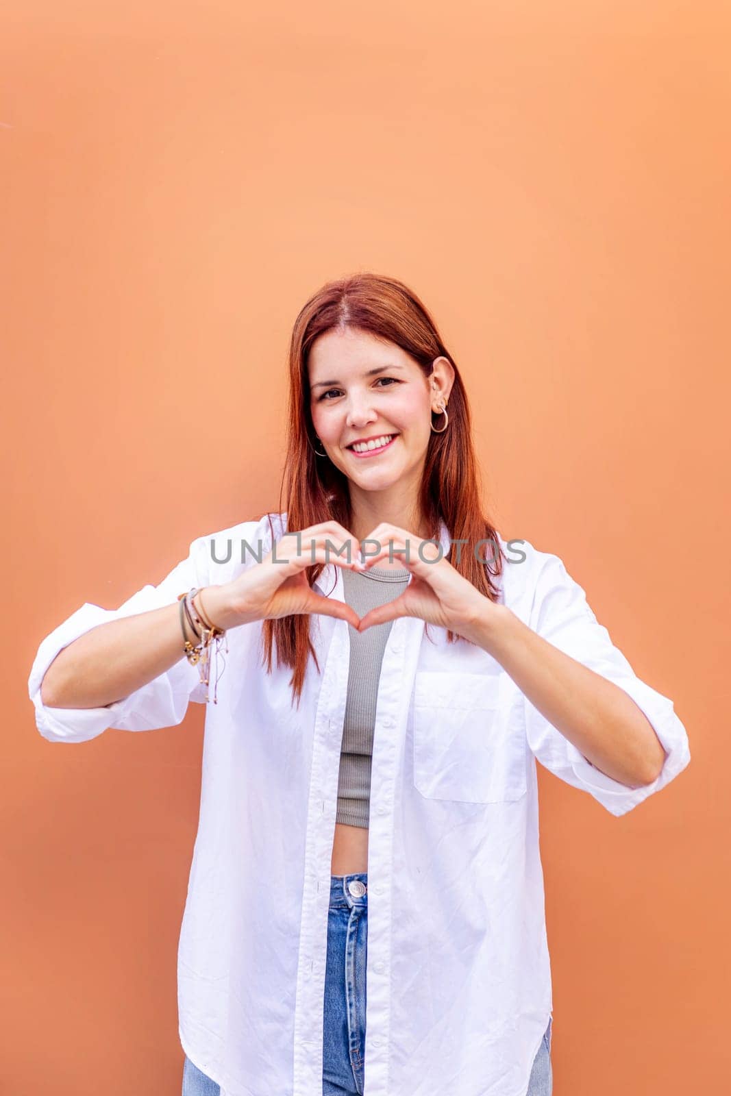 young woman standing in front of orange wall doing a heart shape with hands while looking at camera, she is wearing a white shirt, concept of love, copyspace for text