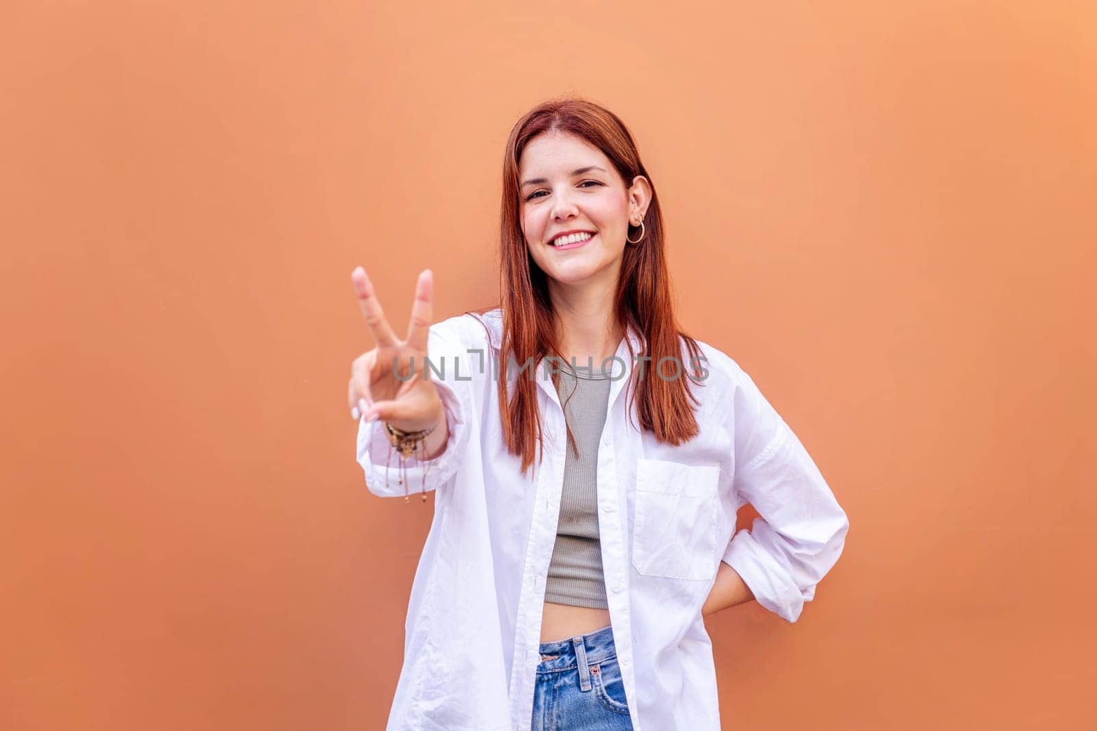 young woman standing in front of orange wall holding up two fingers in victory sign and smiling looking at camera, concept of peace, love and positivity, copyspace for text