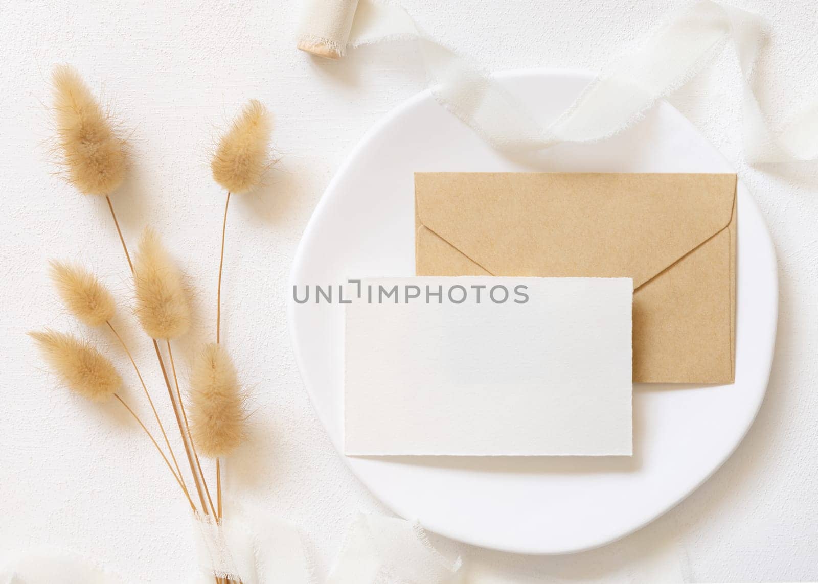 Blank card and envelope on white plate near curled silk ribbons and dried hare's tail grass topview, mockup. Pastel romantic flatlay. Bohemian or close to nature cards for party design or wedding stationery