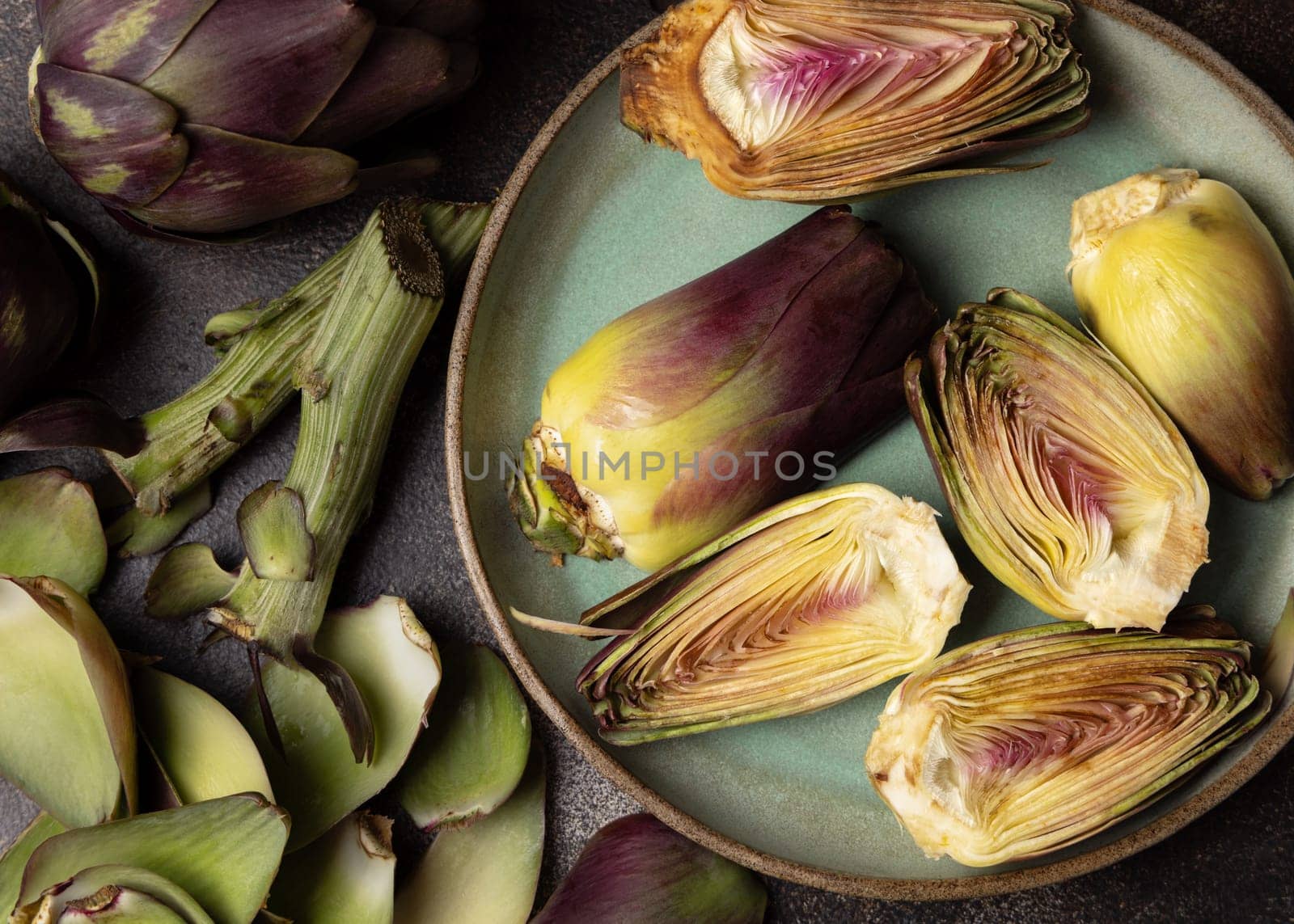 Plate full of cut purple raw romanesco artichokes on dark rustic table top view. Fresh vegetables bought on farmers market. Cleaning and preparing to cook. Italian cousine ingredient