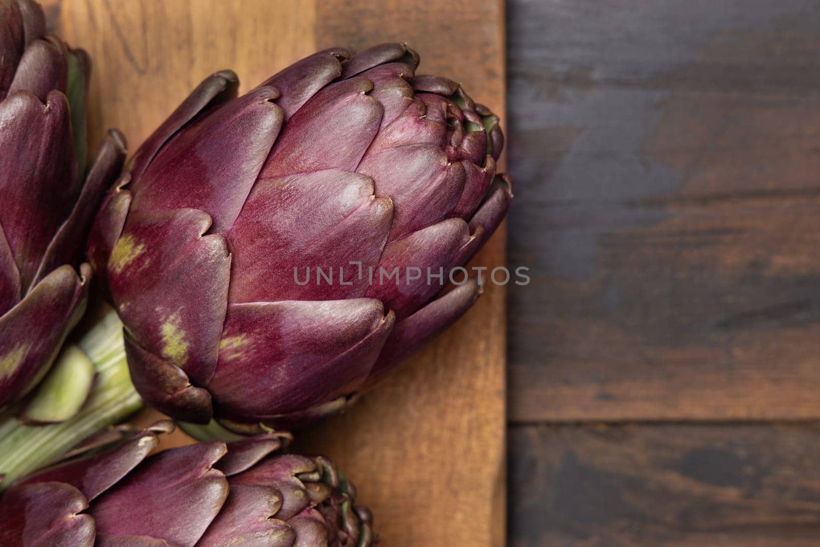 whole purple raw roman artichokes on board on dark weathered wooden table top view, copy space. Fresh vegetables bought on farmers market ready to cook. Italian cousine ingredient