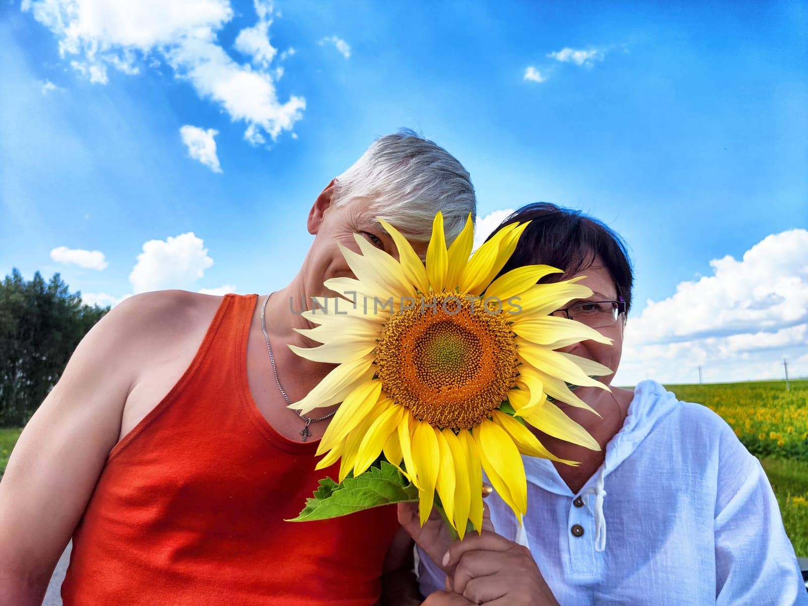 A middle-aged couple is having a joyful moment in the countryside, hiding their smiles behind a large sunflower while enjoying a sunny day during their travels.