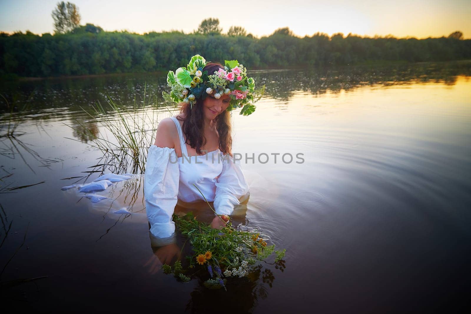 Slavic plump chubby girl in long white dress on the feast of Ivan Kupala with flowers and water in a river or lake on summer evening