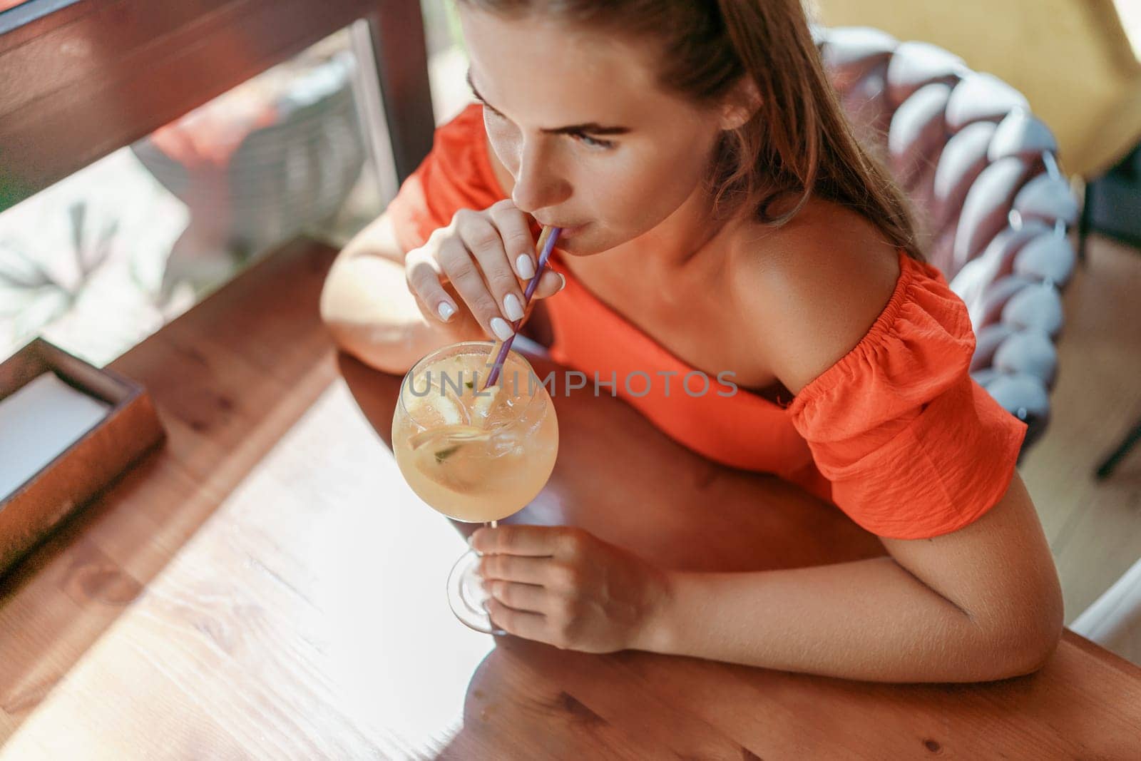 woman in a cafe seated, holding beverage. Dressed in an orange dress with her hair down. Bright interior, large windows allow natural light. Beverage consumption for refreshment