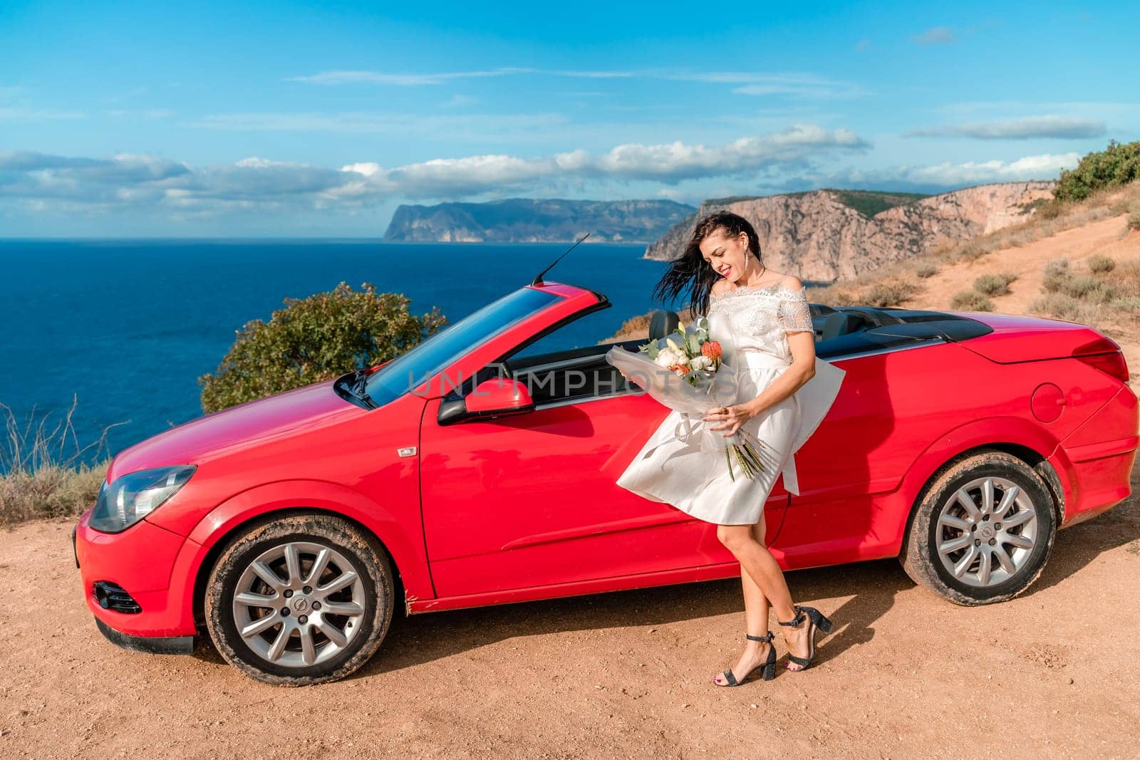 A woman in a white dress stands in front of a red convertible car. She is holding a bouquet of flowers and smiling. Concept of happiness and celebration