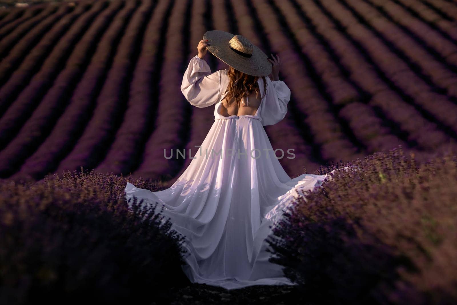 Here's a caption under 150 characters describing the image: Woman in flowing white dress and wide-brimmed hat stands in a vast lavender field at sunset. Rows of purple flowers stretch to the horizon.