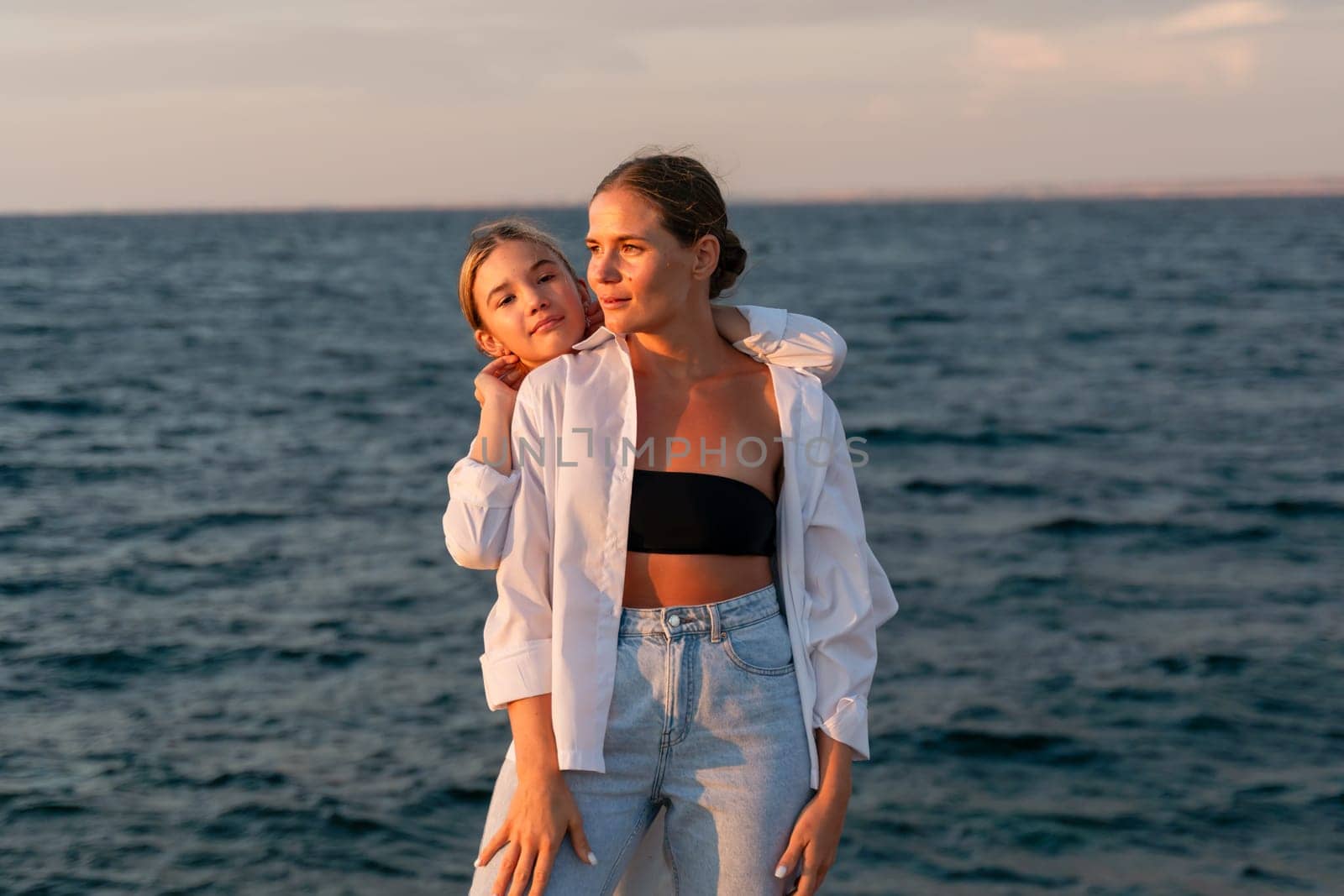 Two women, one in a white shirt and jeans, the other in a black bikini top, stand close together, looking towards the sea at sunset.