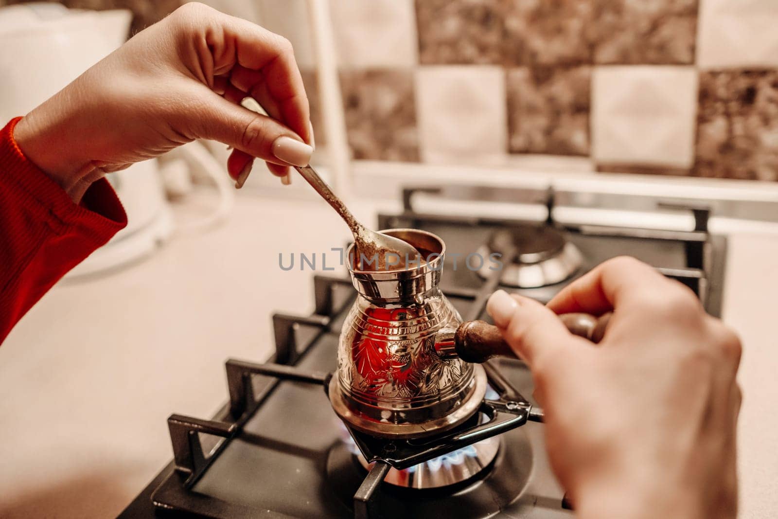 Close-up of hands stirring coffee in a cezve on a gas stove. The metallic cezve sits on a burner, its contents gently mixed.